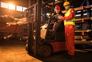 Two workers wearing hard hats and safety gear. One driving an FLT