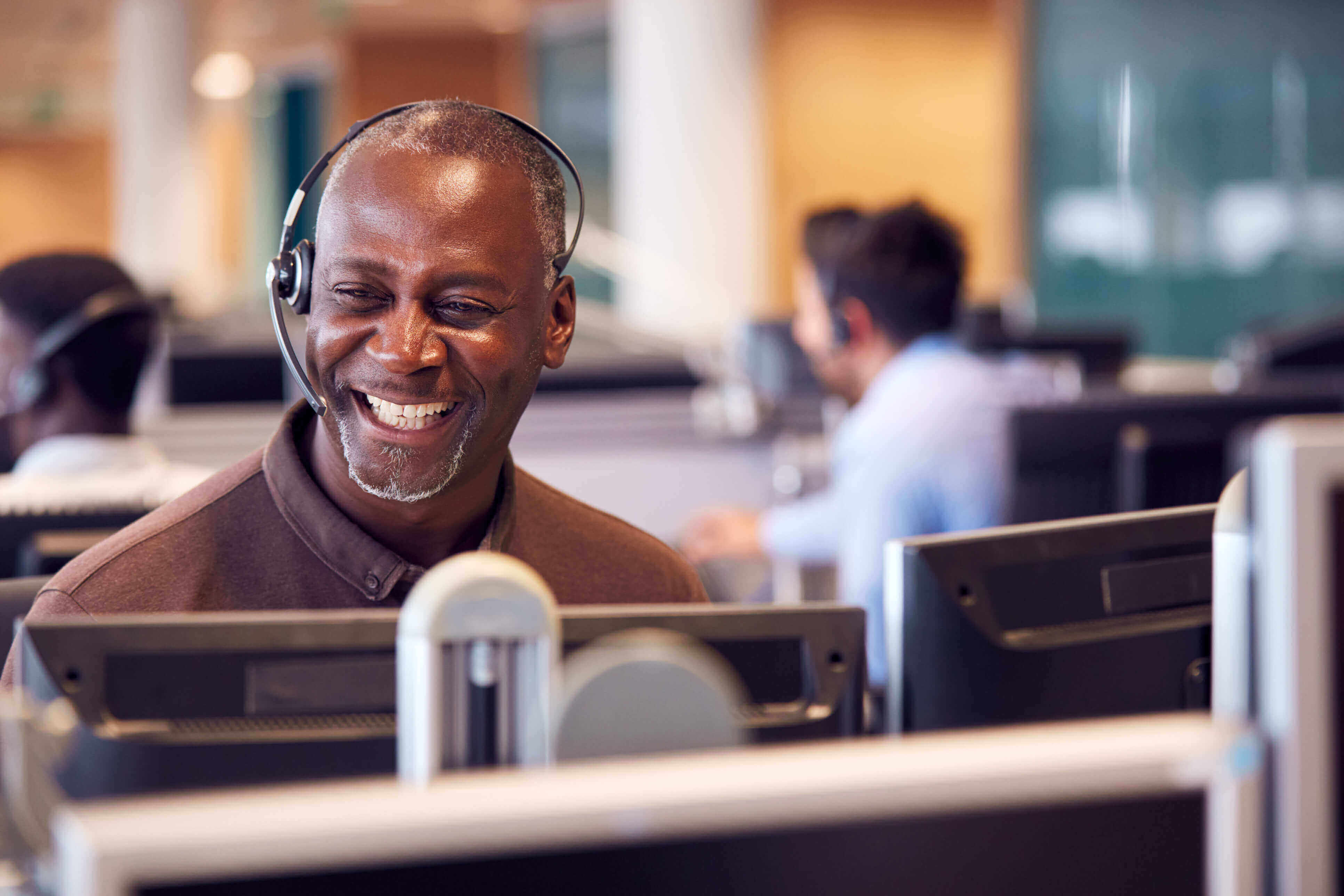 Man wearing a headset, smiling, at a computer in a call centre / telesales department
